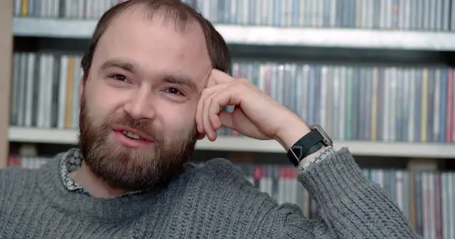 Tomasz Beksiński, zamyślony, z ręką opartą o czoło, na tle regałów z płytami. Jego spojrzenie zdaje się wybiegać poza kadr, jakby rozmyślał o przemijaniu i śmierci.