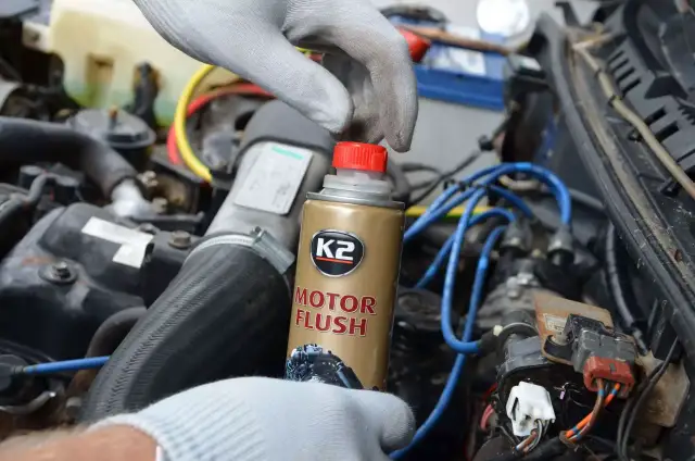Ręka w rękawiczce trzyma puszkę płukanki do silnika K2 Motor Flush na tle silnika samochodowego.