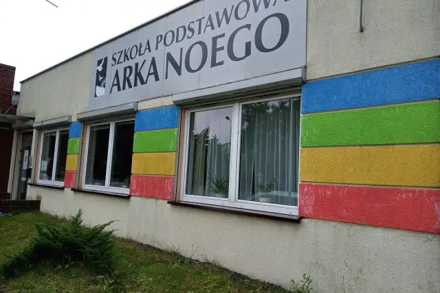 Budynek szkoły z kolorowymi pasami i napisem "SZKOŁA PODSTAWOWA ARKA NOEGO".