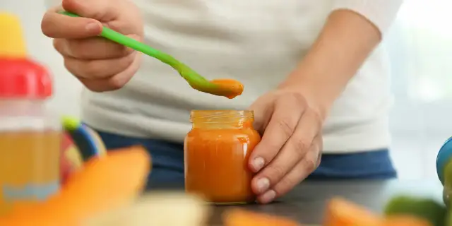 Ręka trzyma zieloną łyżeczkę z pomarańczowym jedzeniem dla niemowląt nad otwartym słoiczkiem.