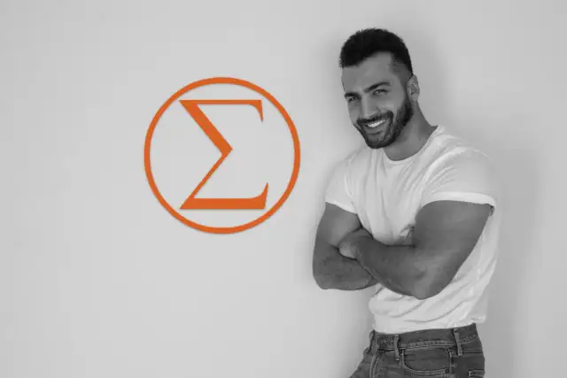 Uśmiechnięty mężczyzna z zarostem, w białej koszulce i jeansach, stoi obok pomarańczowego symbolu Sigma. Jego postawa emanuje pewnością siebie, odzwierciedlając cechy samca sigma.