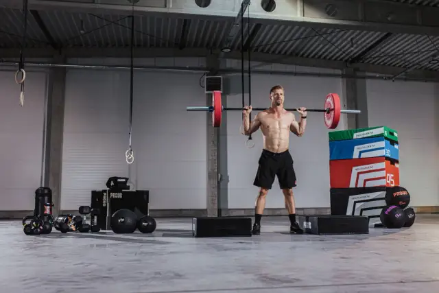 Siłownia pełna sprzętu do treningu. Mężczyzna z idealnym brzuchem podnosi sztangę. To inspiracja, jakie ćwiczenia na brzuch na siłowni wybrać.