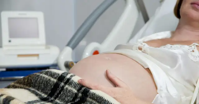 Brzuch kobiety w ciąży, ręka na brzuchu, monitor KTG i łóżko szpitalne.