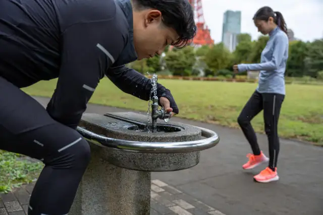 Mężczyzna po bieganiu pije wodę, by złagodzić objawy astmy wysiłkowej. W tle kobieta kontynuuje trening.