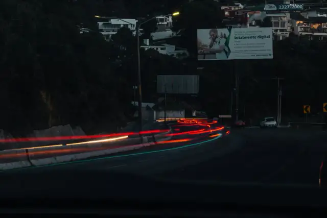 Smugi świateł samochodowych na drodze nocą, z widocznymi budynkami na wzgórzu i billboardem.