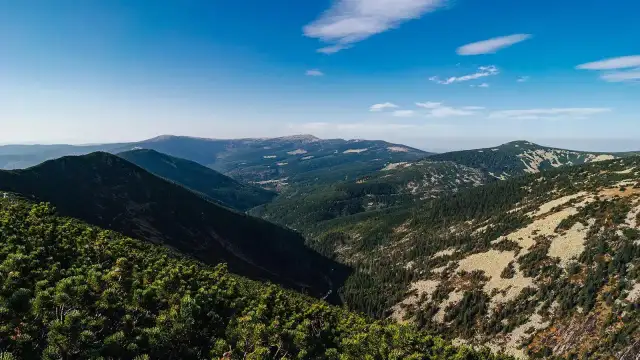 Widok na zielone doliny i skaliste zbocza, gdzie w oddali widać najwyższe szczyty Karkonoszy pod błękitnym niebem.
