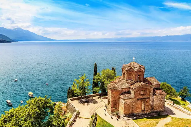 Kamienna cerkiew na wzgórzu nad krystalicznie czystym jeziorem Ochryd. W oddali góry i błękitne niebo.