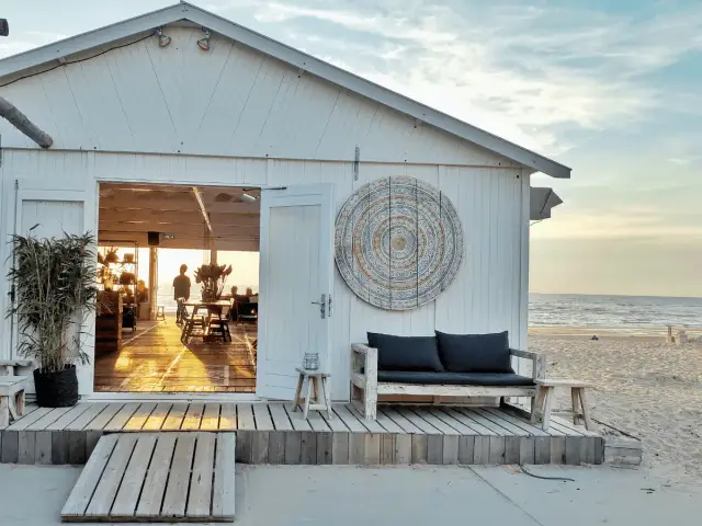 Biały domek plażowy z otwartymi drzwiami, widok na morze i wnętrze z meblami.