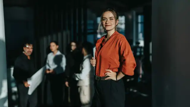 Uśmiechnięta kobieta w pomarańczowej bluzce trzyma teczkę, jakby właśnie odebrała prawo jazdy. W tle grupa osób.