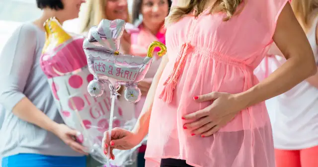 Ciężarna kobieta w różowej sukience trzyma balon w kształcie wózka z napisem "it's a girl!".
