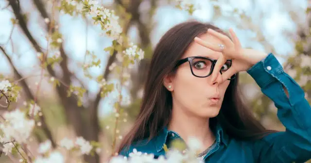 Kobieta w okularach zaciska nos, otoczona kwitnącymi drzewami. Wiosna, piękna, ale dla niej to udręka z powodu kataru siennego.