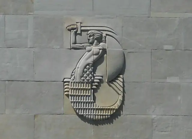 Warszawska Syrenka w płaskorzeźbie na kamiennej ścianie, trzymająca miecz i tarczę.