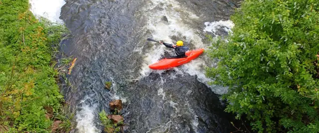 Kajakarz w żółtym kasku płynie czerwonym kajakiem po rwącym nurcie rzeki.