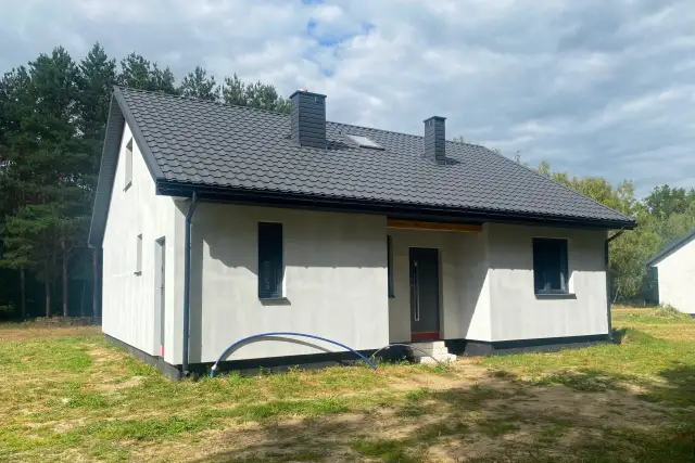 Nowy, biały dom szkieletowy z szarym dachem i dwoma kominami, otoczony zielenią.