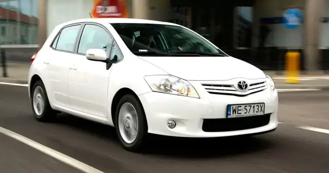 Biała Toyota Auris w ruchu miejskim, z widocznym kierowcą za kierownicą.