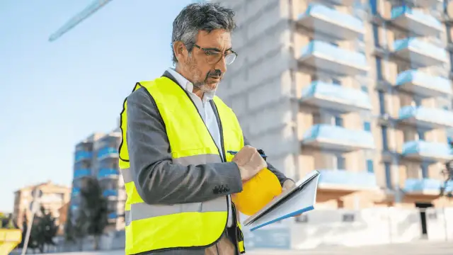 Mężczyzna w kamizelce odblaskowej i kasku trzyma notatnik na tle budowy.
