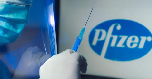 Ręka w rękawiczce trzyma strzykawkę z niebieskim płynem na tle logo Pfizer. Dyskusja o skutkach ubocznych szczepionki Pfizer trwa.