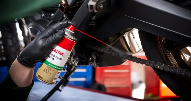 Ręka w czarnej rękawiczce rozpyla smar Castrol na łańcuch motocykla.