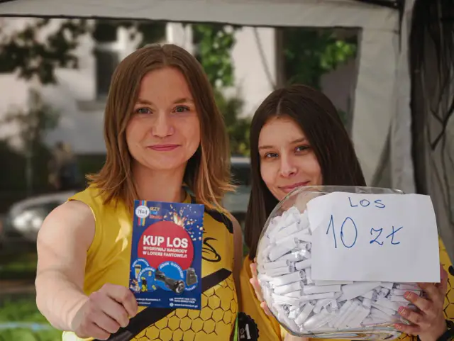 Dwie kobiety w żółtych koszulkach trzymają ulotkę "Kup los" i słoik z losami na loterię fantową.