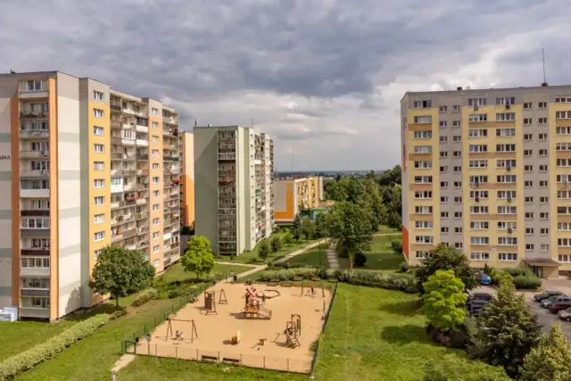 Bloki mieszkalne z placem zabaw na pierwszym planie, pod pochmurnym niebem.