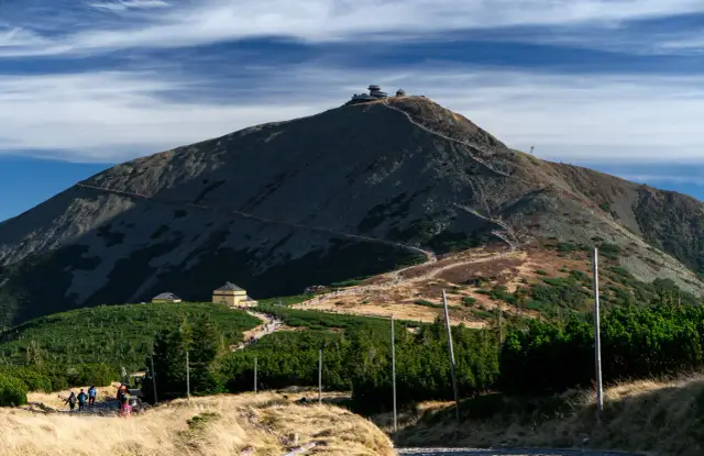Widok na Śnieżkę z łagodnym, kamienistym szlakiem, który wydaje się być najłatwiejszy na zdobycie szczytu. Widać turystów i budynki u podnóża.