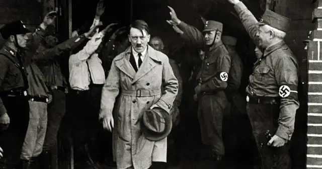 Adolf Hitler w płaszczu, otoczony przez ludzi w mundurach z opaskami ze swastykami, pozdrawiających uniesionymi rękami.