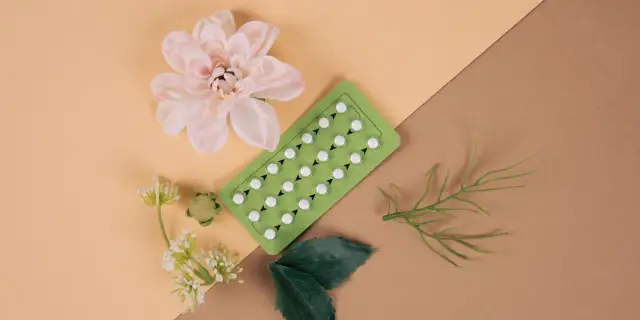 Pigułki antykoncepcyjne na zielonym blistrze, obok kwiatów i liści. Czy od tabletek antykoncepcyjnych się tyje?