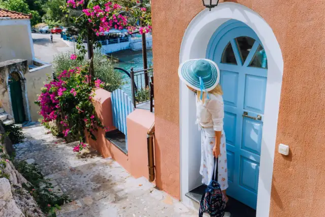 Kobieta w kapeluszu przy niebieskich drzwiach, gotowa na wakacje do Grecji all inclusive. W tle kwitnące bugenwille i morze.
