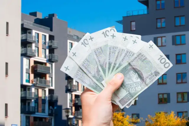 Dłoń trzymająca polskie banknoty 100 zł na tle nowoczesnych budynków mieszkalnych.