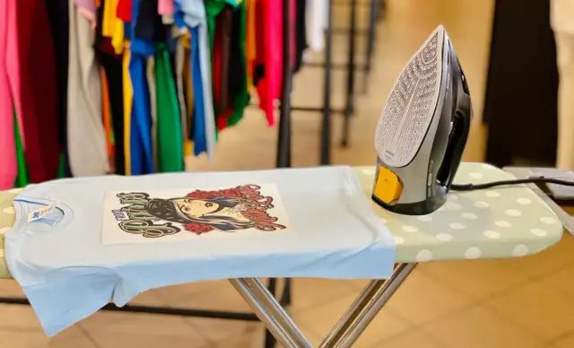 Jak zrobic naprasowanke? Na desce do prasowania leży jasnoniebieska koszulka z nadrukiem, obok żelazko.