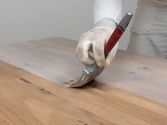 Ręka w rękawiczce nakłada biały preparat na drewniany blat, pokazując, jak usunąć wosk z drewna.