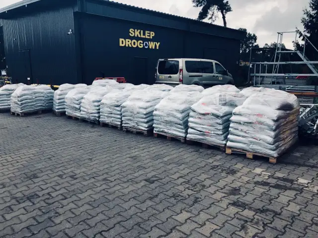 Duże worki z materiałem budowlanym (asfaltem) na paletach przed budynkiem sklepu drogowego.