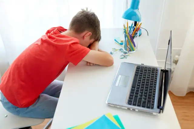 Chłopiec w czerwonej koszulce śpi na biurku obok laptopa i przyborów szkolnych.