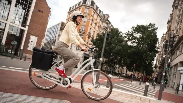Kobieta w kasku jedzie białym rowerem po mieście, z czarną torbą z tyłu.