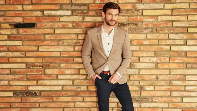 Mężczyzna w beżowej marynarce i granatowych spodniach opiera się o ceglaną ścianę.