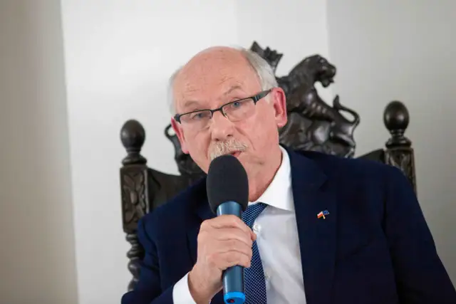 Starszy mężczyzna w okularach i garniturze przemawia do mikrofonu. Na klapie marynarki ma przypinkę z flagami Polski i UE, symbolizującą jego poparcie dla tych, kto głosował za zielonym ładem.