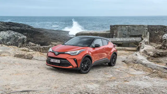 Pomarańczowe, małe auta hybrydowe Toyota C-HR na kamienistym wybrzeżu. W tle widać morze z falami i zniszczony budynek.