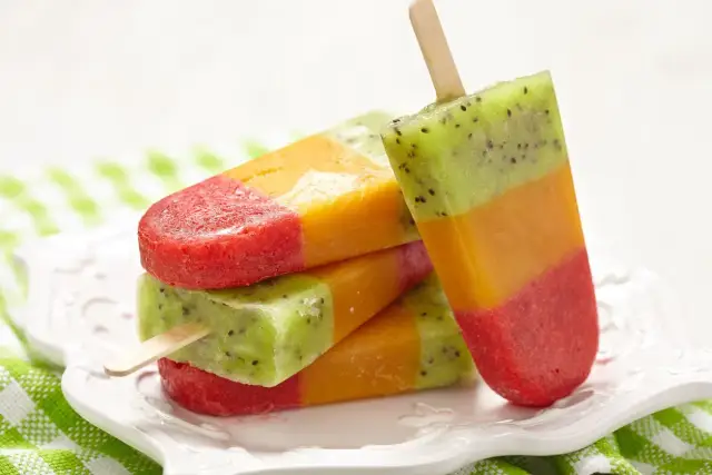 Kolorowe, warstwowe domowe lody na patyku z kiwi, mango i truskawkami, ułożone na białym talerzyku.