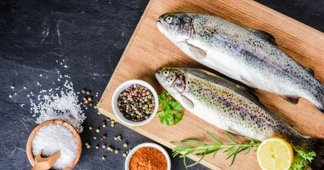 Dwa świeże pstrągi na desce, obok przyprawy i cytryna. Pstrąg to źródło cennych składników odżywczych.
