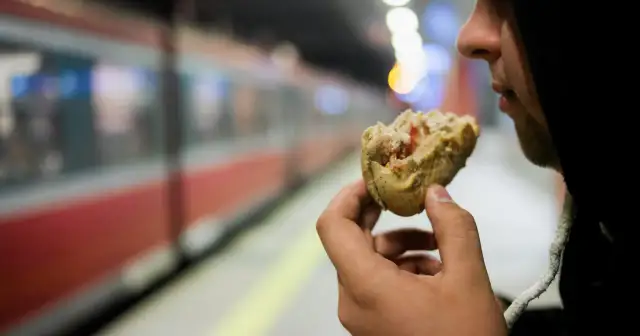 Osoba w kapturze je bułkę na tle pociągu TLK.