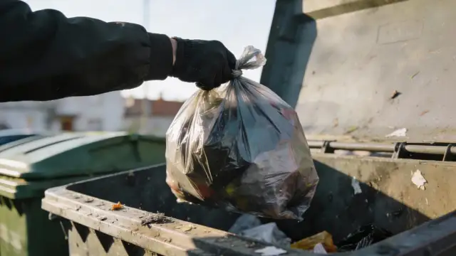 Ręka w czarnej rękawiczce wrzuca worek ze śmieciami do kontenera. Zastanawiamy się, ile zapłacimy za wywóz śmieci.