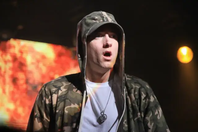 Eminem w bluzie z kapturem i naszyjnikiem, na tle płonącego ekranu.