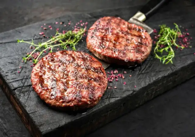 Dwa soczyste, grillowane kotlety na burgery na drewnianej desce z ziołami i różowym pieprzem.