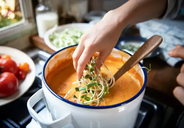 Dłoń dodaje kiełki do pomarańczowej zupy w garnku.