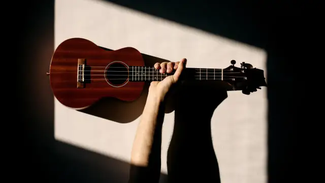 Dłoń trzymająca drewniane ukulele na jasnym tle z geometrycznymi cieniami.