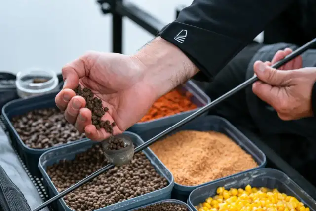 Wędkarz przygotowuje zanętę, trzymając w dłoni pelet. W tle widać pojemniki z różnymi składnikami, w tym kukurydzą.