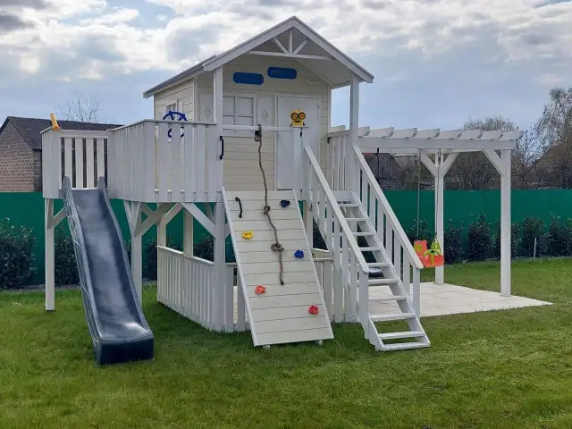 Drewniany domek na placu zabaw z zjeżdżalnią, drabinką, liną i huśtawką.