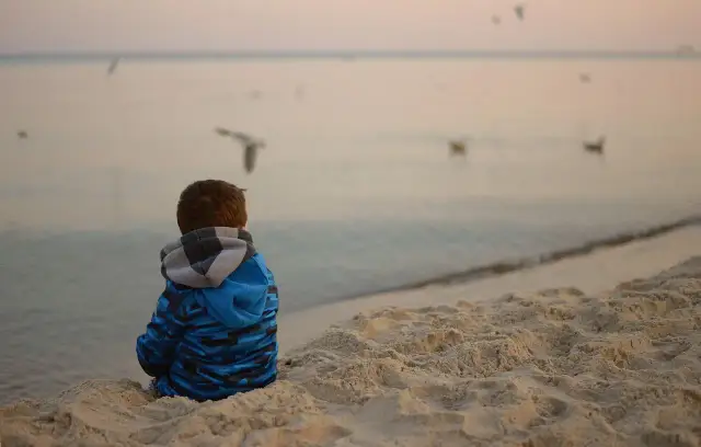 Chłopiec w niebieskiej bluzie siedzi na plaży, obserwując mewy. Spokojna atmosfera nad morzem może pomóc w tym, jak wyciszyć dziecko nadpobudliwe.