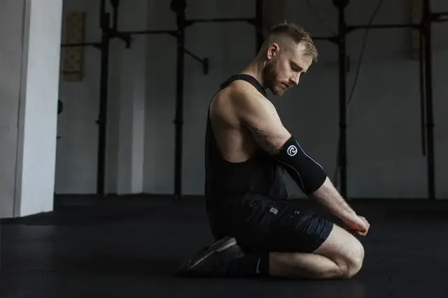 Mężczyzna w czarnym podkoszulku i spodenkach klęczy na siłowni, opierając się na łokciach.