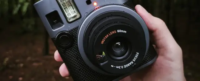 Szary aparat natychmiastowy Instax z obiektywem 60mm, trzymany w dłoni na tle lasu.
